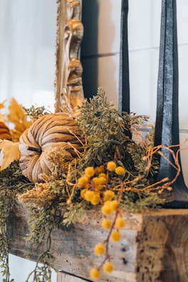 A rustic autumn-themed decoration featuring a pumpkin made of dried leaves and surrounded by various greenery and yellow flowers. Two tall black candles are positioned on a wooden shelf next to an ornate gold mirror frame.