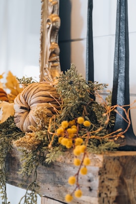 A rustic autumn-themed decoration featuring a pumpkin made of dried leaves and surrounded by various greenery and yellow flowers. Two tall black candles are positioned on a wooden shelf next to an ornate gold mirror frame.