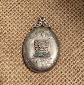 An ornate, oval-shaped silver medal or pendant featuring an engraved design. The central element is a stylized harp surrounded by decorative lines and stars. Beneath the harp, the text reads 'Vancouver' and 'Clean.' The background is a textured burlap material.