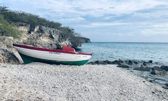 A small boat with the name 'PORTOMARI' is resting on a pebbly beach next to rocky terrain with sparse greenery. The boat is white with green and red stripes. Calm ocean waves gently touch the shore, and the sky is partially cloudy with a hint of light from the setting or rising sun.