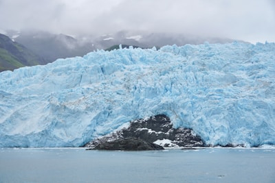 A large glacier with rugged, jagged ice formations stretches across the scene, close to a water body with floating ice pieces. The sky is overcast, contributing to a moody atmosphere, while mountain peaks partially obscured by clouds are visible in the background.