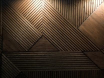 Geometric wooden panel design featuring various rectangular and triangular shapes constructed from lines and planks of wood. The patterns form a cohesive, intricate layout with different angular intersections. Light emphasizes certain sections, creating a warm and textured appearance.