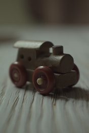 A small, wooden toy train with simple features, including a round chimney and four red wheels, rests on a wooden surface. The lighting is soft and warm, casting gentle shadows and creating a vintage ambiance.