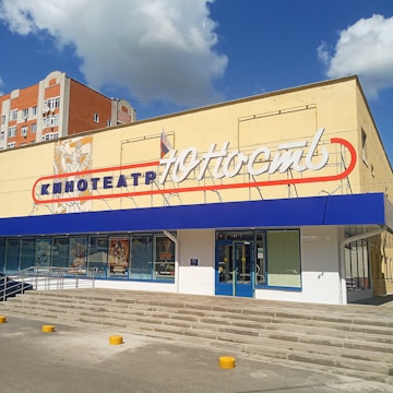 A building with a cinema sign written in Cyrillic characters, featuring a blue awning and poster displays in large windows. The building has light-colored walls, and a set of stairs leads up to the entrance. A red and white sign adorns the upper part of the facade, with additional signage below.
