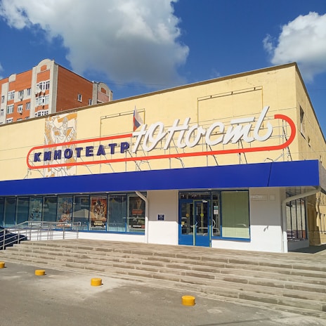 A building with a cinema sign written in Cyrillic characters, featuring a blue awning and poster displays in large windows. The building has light-colored walls, and a set of stairs leads up to the entrance. A red and white sign adorns the upper part of the facade, with additional signage below.