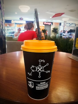 A black takeaway coffee cup with a yellow lid sits on a wooden table in a cafe. The cup features a caffeine molecule diagram and Russian text. In the background, a cafe environment with blurred individuals chatting.