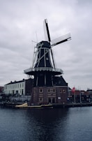 A traditional Dutch windmill stands prominently by a canal, surrounded by brick and white facade buildings. The sky is overcast, adding a somber tone to the scene. A small boat is docked in the canal nearby.