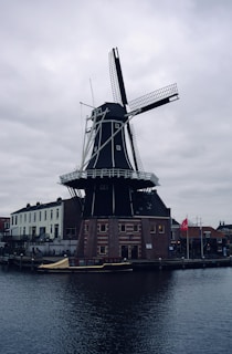 A traditional Dutch windmill stands prominently by a canal, surrounded by brick and white facade buildings. The sky is overcast, adding a somber tone to the scene. A small boat is docked in the canal nearby.