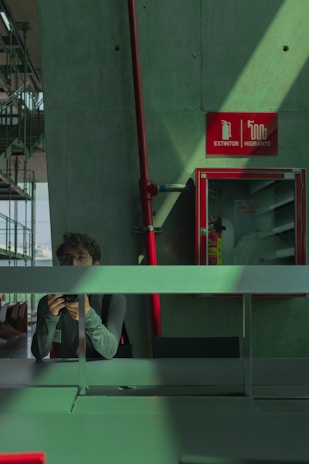 A person is reflected in a glass surface, holding a camera and taking a photo indoors. There is a concrete wall adorned with a red fire extinguisher cabinet and safety signage. The lighting casts shadows, creating a dynamic pattern on the green and red surfaces.