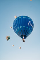 A group of colorful hot air balloons ascends into a clear blue sky, with one prominent blue balloon in the foreground displaying some text. Smaller balloons are scattered throughout the background, creating a sense of depth and activity.