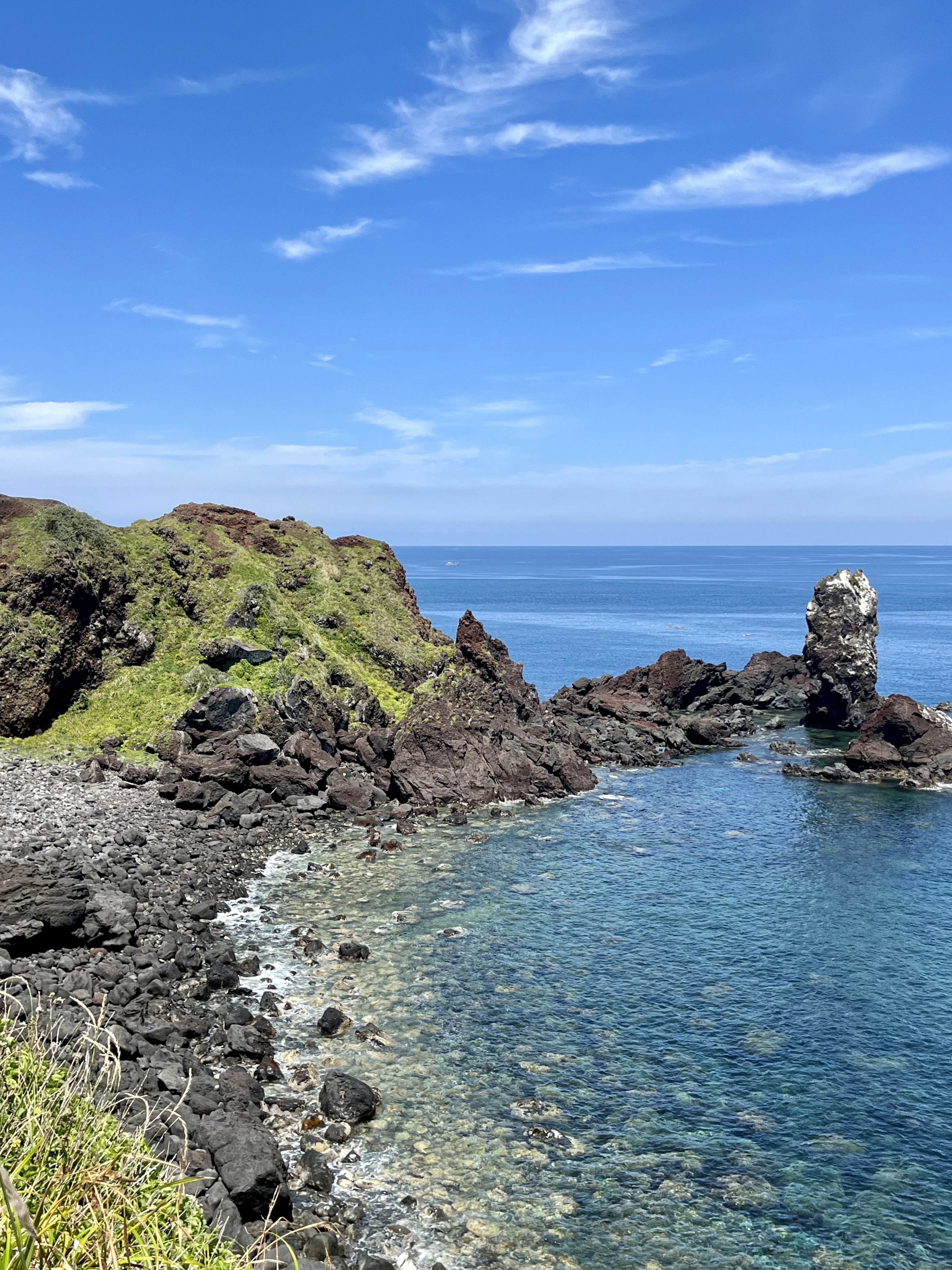 A jagged lava sea stack rises from clear turquoise water beside a rocky black coastline.