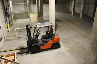 A forklift is parked indoors in a concrete industrial setting, with an empty wooden crate on the ground nearby. The walls are made of cinderblocks, and there are yellow direction arrows on the concrete floor.