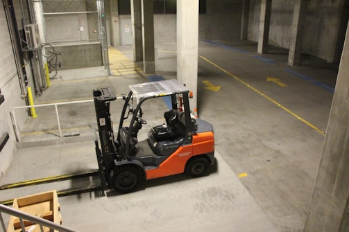 A forklift is parked indoors in a concrete industrial setting, with an empty wooden crate on the ground nearby. The walls are made of cinderblocks, and there are yellow direction arrows on the concrete floor.