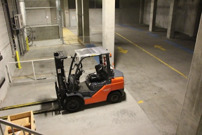 A forklift is parked indoors in a concrete industrial setting, with an empty wooden crate on the ground nearby. The walls are made of cinderblocks, and there are yellow direction arrows on the concrete floor.
