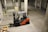 A forklift is parked indoors in a concrete industrial setting, with an empty wooden crate on the ground nearby. The walls are made of cinderblocks, and there are yellow direction arrows on the concrete floor.