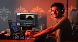 A person wearing headphones is seated at a desk with multiple monitors displaying editing software. The workspace includes a camera, a clock with LED display, and a cup containing pens. The person is smiling and looking at the camera, creating a friendly and engaging atmosphere.