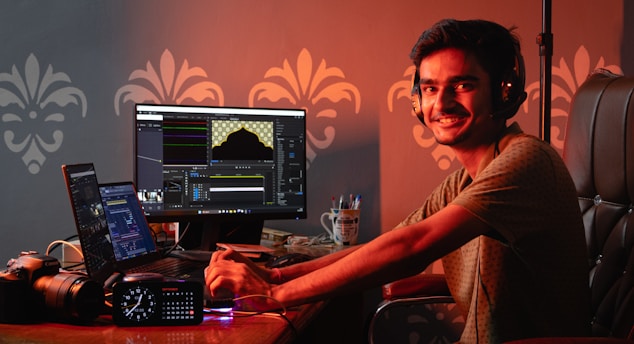 A person wearing headphones is seated at a desk with multiple monitors displaying editing software. The workspace includes a camera, a clock with LED display, and a cup containing pens. The person is smiling and looking at the camera, creating a friendly and engaging atmosphere.