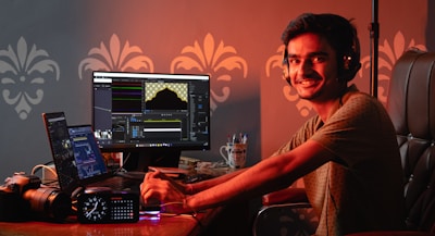 A person wearing headphones is seated at a desk with multiple monitors displaying editing software. The workspace includes a camera, a clock with LED display, and a cup containing pens. The person is smiling and looking at the camera, creating a friendly and engaging atmosphere.
