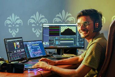 Smiling Indian video editor with headphones working on a laptop and monitor
