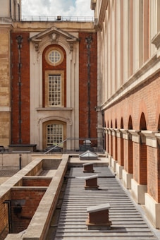 The image depicts a perspective view of a historical brick building with classical architectural features, including large arched windows and decorative stone elements. The building is flanked by stone columns, and the scene includes a roof with small structures protruding from it.