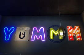 A vibrant sign spells out the word 'YUMM' using various styles of illuminated and neon letters. The first letter 'Y' is in bright blue neon. The 'U' resembles a road sign with the word 'COUNTY' above it, showing a worn-out metallic finish. The first 'M' is pink with a neon glow, and the second 'M' is yellow with a hanging light fixture in front of it, giving a unique visual effect. The final 'M' is red with illuminated white dots.