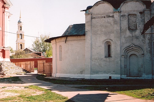 A historic architectural scene featuring an old white stone building with arched windows, adjacent to a red brick building with steps. In the background, a tall bell tower with a golden spire is visible. The area is surrounded by wooden fences, and there is a path leading from the building to the gate.