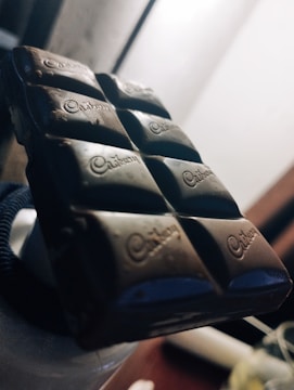 A close-up view of a dark chocolate bar with the brand name embossed on each square segment. The lighting highlights the bar's smooth texture and dark color, with a soft background blur.