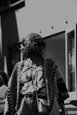 A person in a patterned, oversized cardigan and a printed blouse, wearing sunglasses and a headscarf, stands in an urban setting. The photo has a black and white filter, creating a classic, timeless feel.