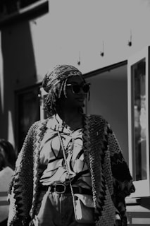 A person in a patterned, oversized cardigan and a printed blouse, wearing sunglasses and a headscarf, stands in an urban setting. The photo has a black and white filter, creating a classic, timeless feel.