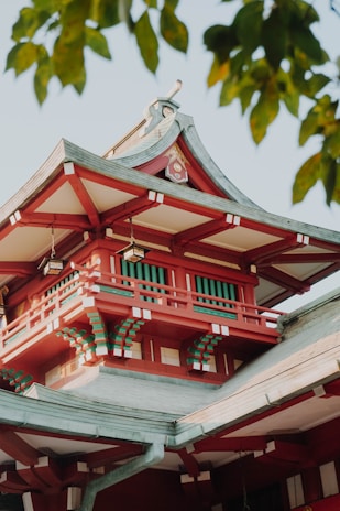 A traditional Japanese architectural style visible through the top section of a building. The structure features intricate wooden craftsmanship, highlighted by vibrant red and subtle green accents. The roof is elegantly curved and adorned with decorative elements. Sunlight illuminates the building, and green leaves frame the scene, suggesting a tranquil, natural setting.