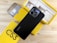 A smartphone with a black back featuring dual camera lenses is placed atop a partially open yellow box. The box is labeled 'C53' and contains various accessories, such as a charger and cables. A protective phone case is also visible beside the box.