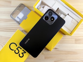 A smartphone with a black back featuring dual camera lenses is placed atop a partially open yellow box. The box is labeled 'C53' and contains various accessories, such as a charger and cables. A protective phone case is also visible beside the box.