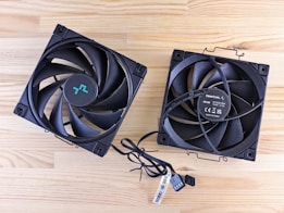 Two black computer cooling fans are placed on a light wooden surface. The fans have multiple blades and visible wires. One fan displays branding and specifications on the central hub.
