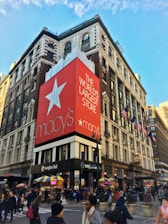 A bustling urban street corner features a large department store building with a prominent red banner displaying white text that reads 'The World's Largest Store.' The store's logo is visible. People are walking along the sidewalks, and there are yellow and blue awnings over small vendor stalls. American flags are mounted along the building façade, and the architecture includes classic features with large windows.