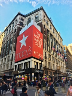 A bustling urban street corner features a large department store building with a prominent red banner displaying white text that reads 'The World's Largest Store.' The store's logo is visible. People are walking along the sidewalks, and there are yellow and blue awnings over small vendor stalls. American flags are mounted along the building façade, and the architecture includes classic features with large windows.