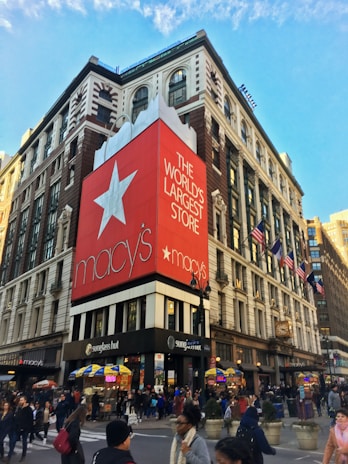 A bustling urban street corner features a large department store building with a prominent red banner displaying white text that reads 'The World's Largest Store.' The store's logo is visible. People are walking along the sidewalks, and there are yellow and blue awnings over small vendor stalls. American flags are mounted along the building façade, and the architecture includes classic features with large windows.
