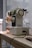 A sewing machine is placed on a wooden table, with various knobs and dials visible on its front panel. The machine appears to be an older model with a white and beige color scheme. It is plugged into a power outlet, and there are some sewing accessories and tools nearby. The background consists of a tiled wall and a window, allowing natural light to illuminate the scene.