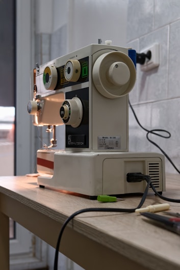 A sewing machine is placed on a wooden table, with various knobs and dials visible on its front panel. The machine appears to be an older model with a white and beige color scheme. It is plugged into a power outlet, and there are some sewing accessories and tools nearby. The background consists of a tiled wall and a window, allowing natural light to illuminate the scene.
