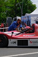 A man wearing a blue shirt works on a red and white race car that has logos like 'SUPERBET' and 'UniCredit Leasing' on it. The car is outdoors, near a setup that includes a tent and a sign with 'ONELIFT'. Green trees and a parked van are visible in the background.