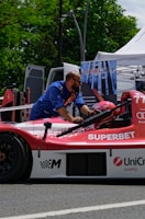A man wearing a blue shirt works on a red and white race car that has logos like 'SUPERBET' and 'UniCredit Leasing' on it. The car is outdoors, near a setup that includes a tent and a sign with 'ONELIFT'. Green trees and a parked van are visible in the background.