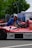 A man wearing a blue shirt works on a red and white race car that has logos like 'SUPERBET' and 'UniCredit Leasing' on it. The car is outdoors, near a setup that includes a tent and a sign with 'ONELIFT'. Green trees and a parked van are visible in the background.