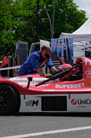 A man wearing a blue shirt works on a red and white race car that has logos like 'SUPERBET' and 'UniCredit Leasing' on it. The car is outdoors, near a setup that includes a tent and a sign with 'ONELIFT'. Green trees and a parked van are visible in the background.