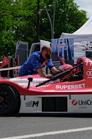 A man wearing a blue shirt works on a red and white race car that has logos like 'SUPERBET' and 'UniCredit Leasing' on it. The car is outdoors, near a setup that includes a tent and a sign with 'ONELIFT'. Green trees and a parked van are visible in the background.