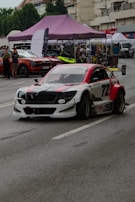A modified racing car with a distinctive red, white, and black color scheme is driving on a street, likely participating in a motorsport event. The car features aerodynamic enhancements such as a rear wing and custom bodywork. In the background, spectators are gathered under tents with other cars on display. The scene appears to be set in an urban environment with buildings and trees visible.