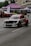 A modified racing car with a distinctive red, white, and black color scheme is driving on a street, likely participating in a motorsport event. The car features aerodynamic enhancements such as a rear wing and custom bodywork. In the background, spectators are gathered under tents with other cars on display. The scene appears to be set in an urban environment with buildings and trees visible.