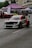 A modified racing car with a distinctive red, white, and black color scheme is driving on a street, likely participating in a motorsport event. The car features aerodynamic enhancements such as a rear wing and custom bodywork. In the background, spectators are gathered under tents with other cars on display. The scene appears to be set in an urban environment with buildings and trees visible.
