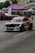 A modified racing car with a distinctive red, white, and black color scheme is driving on a street, likely participating in a motorsport event. The car features aerodynamic enhancements such as a rear wing and custom bodywork. In the background, spectators are gathered under tents with other cars on display. The scene appears to be set in an urban environment with buildings and trees visible.