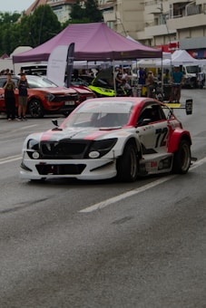 A modified racing car with a distinctive red, white, and black color scheme is driving on a street, likely participating in a motorsport event. The car features aerodynamic enhancements such as a rear wing and custom bodywork. In the background, spectators are gathered under tents with other cars on display. The scene appears to be set in an urban environment with buildings and trees visible.