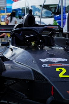 A sleek black racing car is positioned in an outdoor setting, featuring aerodynamic design and carbon fiber material. A bright number '2' is visible on the body, with various stickers and logos. In the background, vans and blurred figures suggest a busy environment, possibly a racetrack or event area.