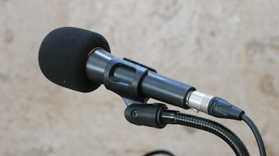 A black microphone with a windscreen cover is mounted on a stand against a blurred light brown background. The microphone has an attached cable visible in the foreground.